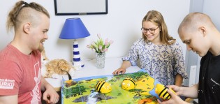 Kaksi nuorta miesoletettua ja yksi naisoletettua testaavat pöydällä olevaa mehiläispeliä hymy huulillaan.