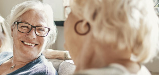 Kaksi iäkkäämpää naista istuu sohvalla vierekkäin ja katsovat toisiinsa hymyillen.
