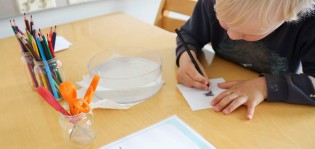 Kuvassa lapsi piirtää paperille, edessä kyniä ja saksia kipoissa.
