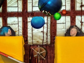 kaksi miehen päätä pilkistää keltaisten patjojen alta tehdasmiljöössä, mukana sirkusvälineitä