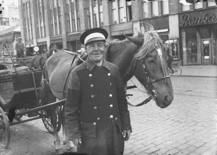 Mustavalkoisessa kuvassa kuljettaja hymyilee hevoskärryn kanssa.