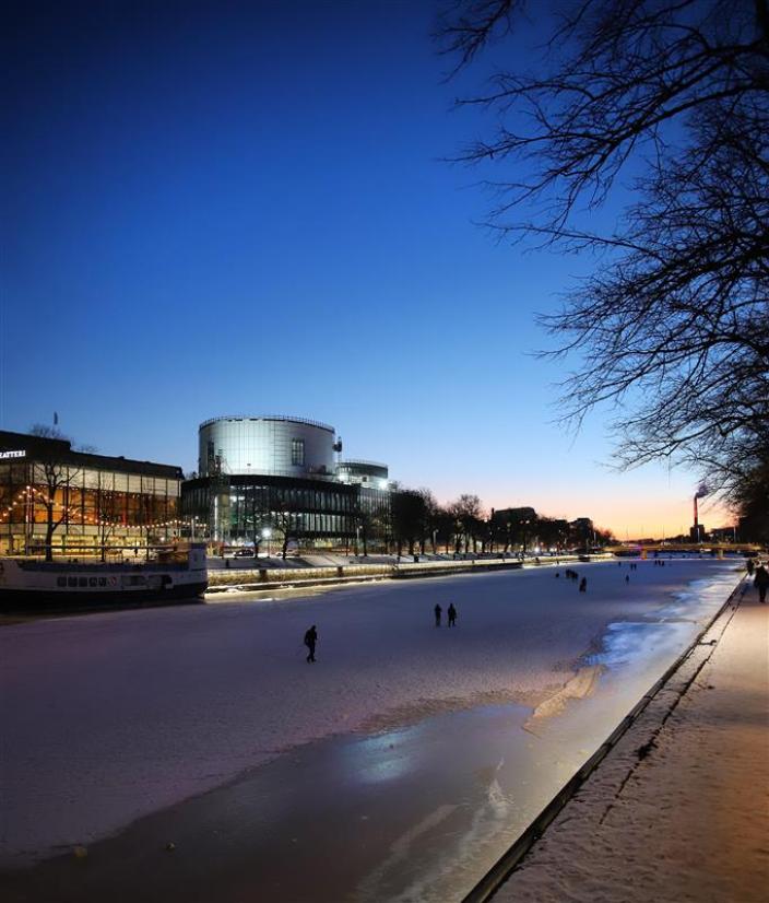 Jäätyneen joen rannalla iltavalaistuksessaan loistavat Turun musiikkitalo Fuuga ja Turun Kaupunginteatteri. Joen jäällä on kävelemässä joitakin ihmisiä auringon laskiessa. (Kuva: Marko Kivistö)