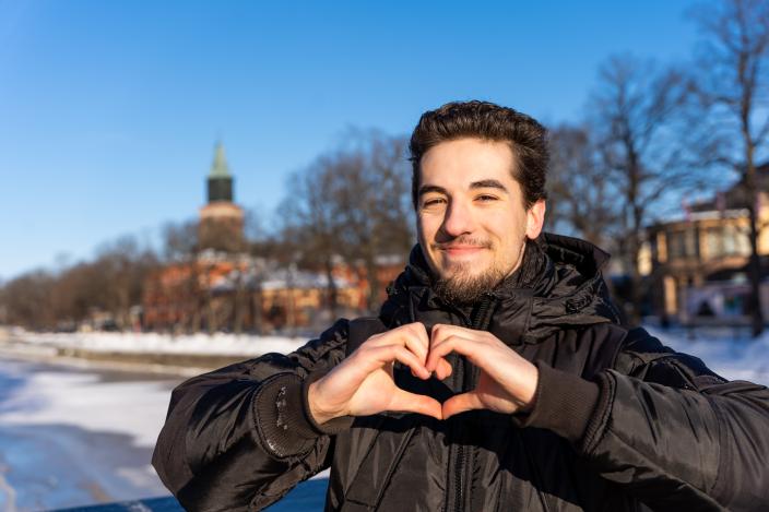Sillalla seisova opiskelija muodostaa käsillään sydämen. Taustalla jäätynyt Aurajoki ja Tuomiokirkko. (Foto: Hanna Oksanen)
