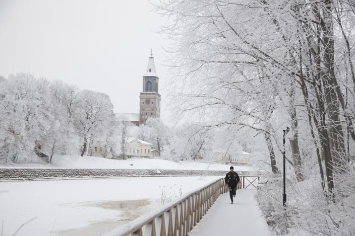 Kuva talvisesta jokirannasta, näkymä Tuomiokirkkoa kohden, etualalla kävelijä.