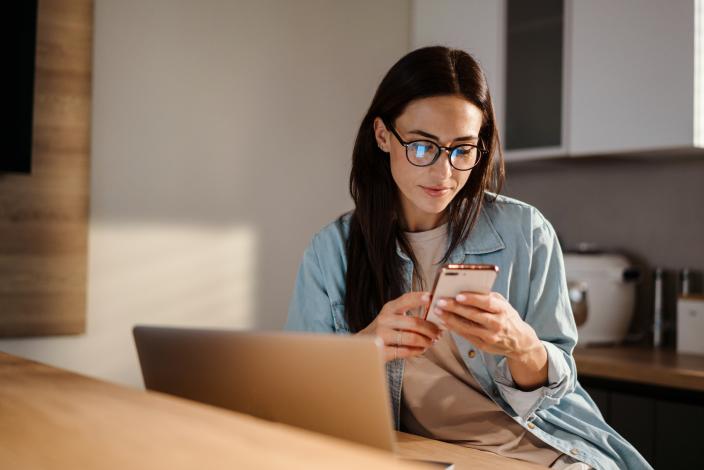 Nainen selaamassa puhelinta samalla, kun hän työskentelee tietokoneella. (Picture: Drobot Dean/Adobe Stock)