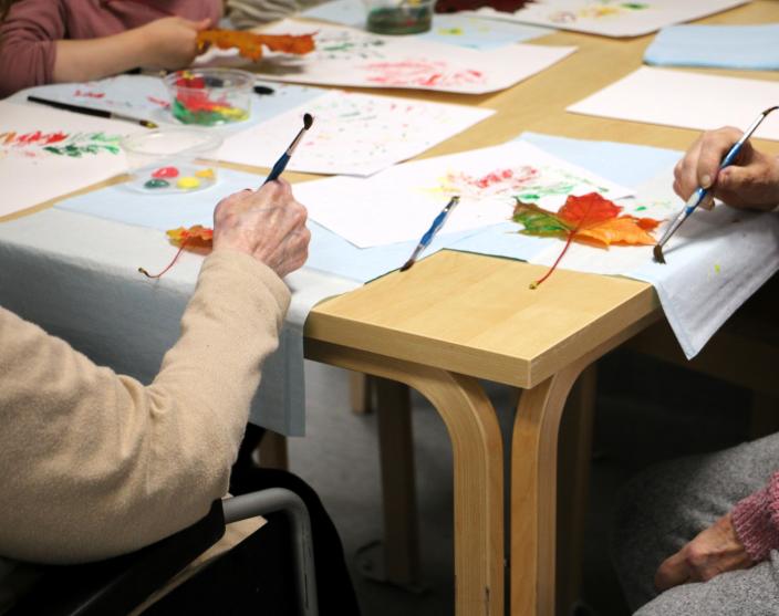 Händerna på två äldre människor och ett barn som målar höstlöven. (Foto: Hannele Sivonen)