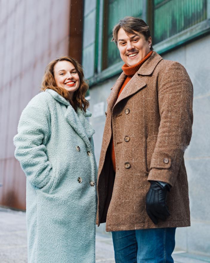 Kaksi henkilöä seisoo ulkona ison rakennuksen seinustan edessä ja katsoo hymyillen kameraan. (Kuva: Heikki Räisänen)