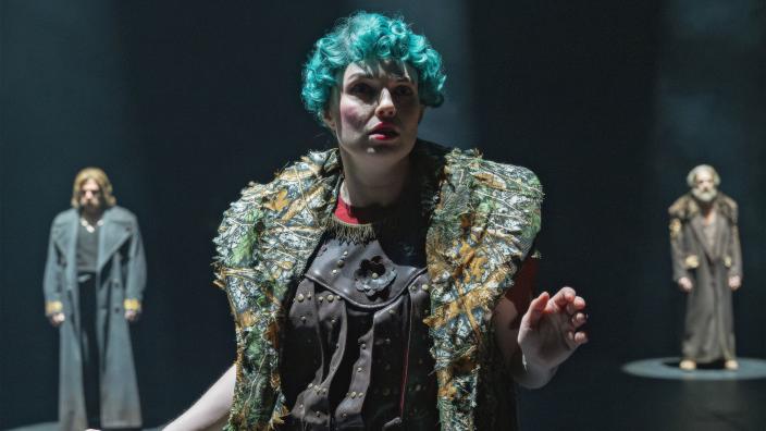 Rooliasuun pukeutunut henkilö seisoo estradilla. Hänen takanaan molemmilla puolilla seisoo rooliasuinen henkilö. (Kuva: Otto-Ville Väätäinen)