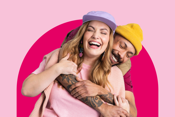 Kaksi ihmistä halaavaat nauraen, vaaleanpunainen tausta.