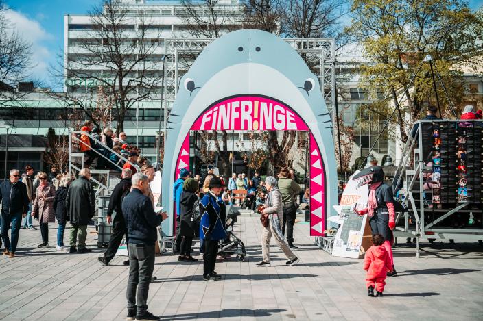 Ihmisiä kulkee ja kokoontuu Finfringe-festivaalin portin ympärille Turun Kauppatorilla. Etualalla näkyy maskottihahmo ja lapsi, taustalla katsomorakenteita ja esitystaulu, joka esittää haita. (Kuva: Finfringe)
