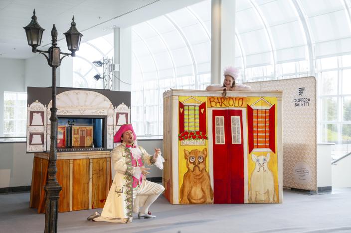 teatterilavasteiden edessä hurmaavasti pukeutunut mies polvillaan hattu päässä (Foto: Matilda Gronow, Suomen kansallisooppera)