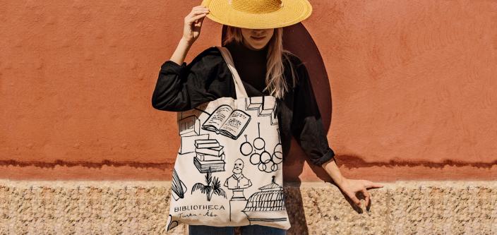 Ihminen nojaa seinään, hellehattu peittää kasvot,Bibliotheca-kangaskassi on olkapäällä  (Kuva: Olli Sallinen)