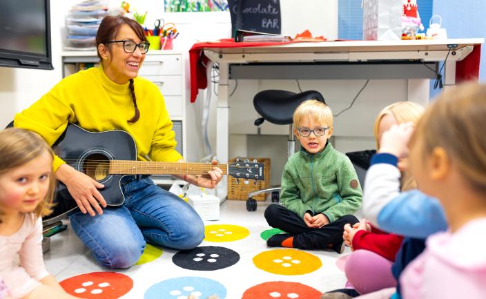 En vuxen sitter på golvet och små barn sitter i en ring framför henne. Den vuxna har en gitarr i famnen. (Foto: Heikki Räisänen)