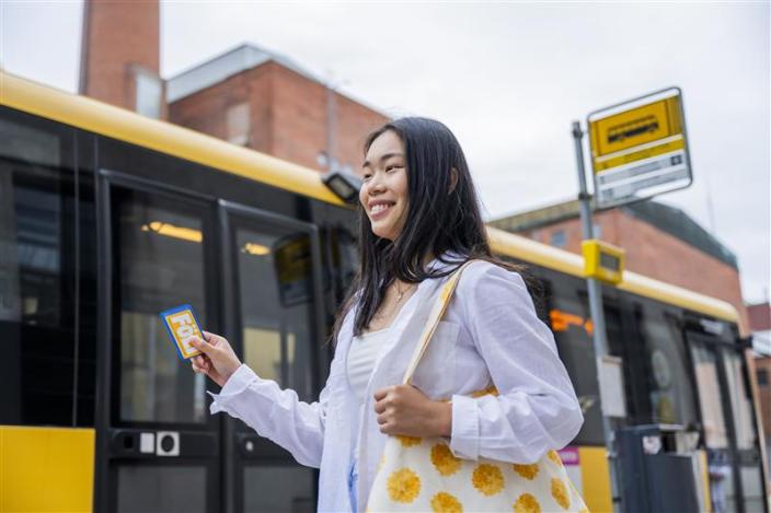 Nainen seisoo bussipysäkillä keltaisen Föli-bussin vieressä, hänellä on kädessään bussikortti. (Foto: Vesa Tyni)