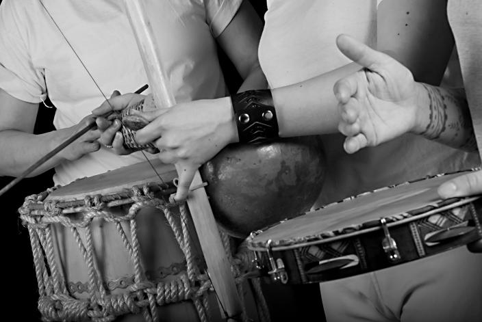 Mustavalkoinen valokuva, jossa kolme henkilöä soittaa erilaisia rumpuja. Kuva on rajattu niin, että henkilöiden kasvot eivät näy. (Kuva: Capoeiraskolan Senzala)