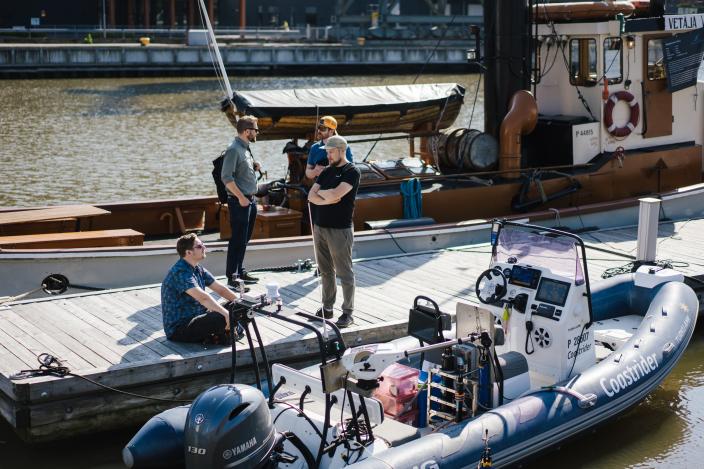 vene kiinnitettynä laituriin. Neljä henkilöä juttelee laiturilla. Taustalla joki. (Kuva: Tara Jaakkola)