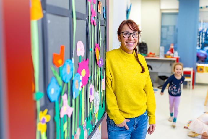 En vuxen står bredvid väggen på dagis och tittar på kameran med ett leende. I bakgrunden springer ett barn mot den vuxna. (Foto: Heikki Räisänen)