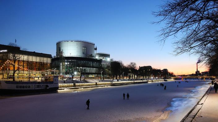 Åbo Stadsteater och det byggande Åbo musikhus Fuga är upplysta vid den frusna Aura å. Några människor promenerar på isen när solen går ner. (Foto: Marko Kivistö)
