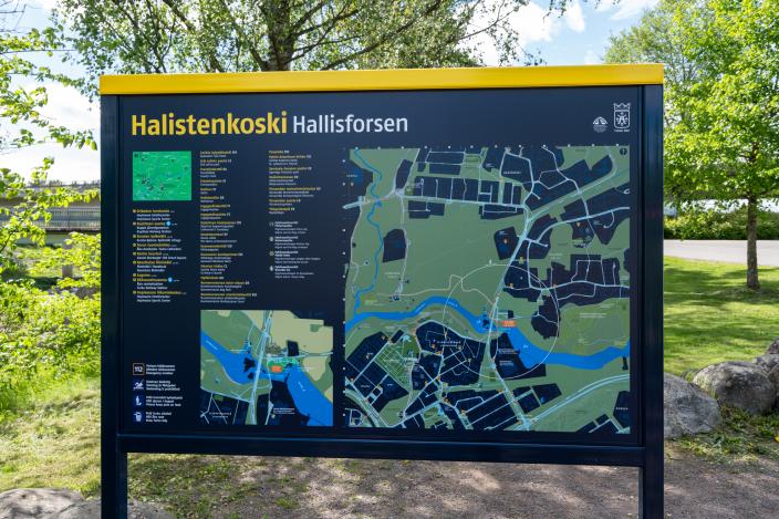 Halistenkoskesta kertova musta opastekyltti, jossa on kartta ja tekstiä. Taustalla on kesäistä nurmikkoa ja puita. (Kuva: Focus-Video)