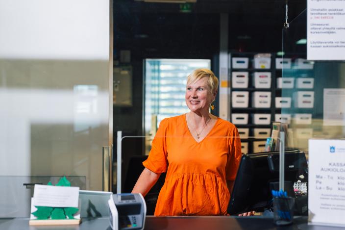 Henkilö seisoo asiakaspalvelutiskin takana ja hymyilee (Kuva: Heikki Räisänen)