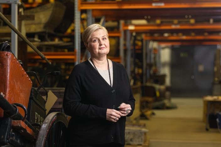 Vaaleahiuksinen nainen seisoo esinevarastossa ja katsoo kameraan. (Kuva: Ville Mäkilä)