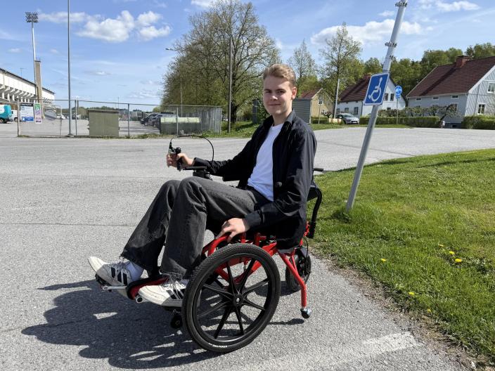 En hyrbar rullstol för anpassad motion i utomhusmiljö. En person sitter i rullstolen och använder en elektrisk styrspak på en asfalterad väg. (Foto: Johanna Friman)