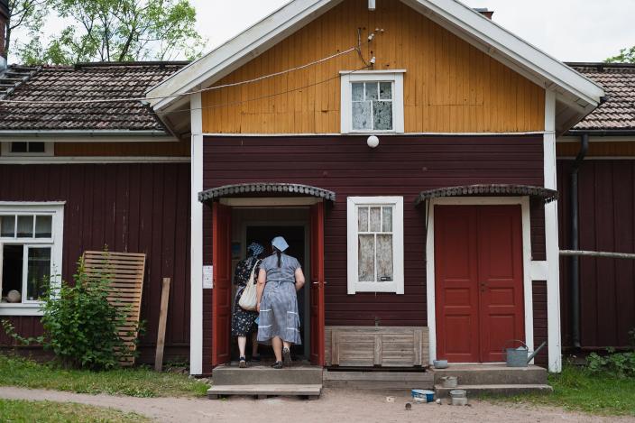 Kaksi 50-luvun emännän asuun pukeutunutta henkilöä kävelee vanhan maatalon pääovesta sisään. On kesä. (Kuva: Joonas Mäkivirta)