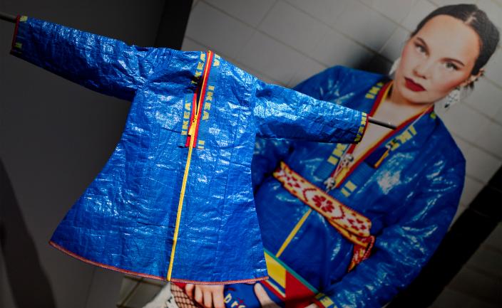 Etualalla on Ikean sinisistä kasseista tehty pienikokoinen saamelaispuku. Samantapaiseen kolttaan pukeutunut henkilö näkyy taustakuvassa. (Kuva: Mika Friman)