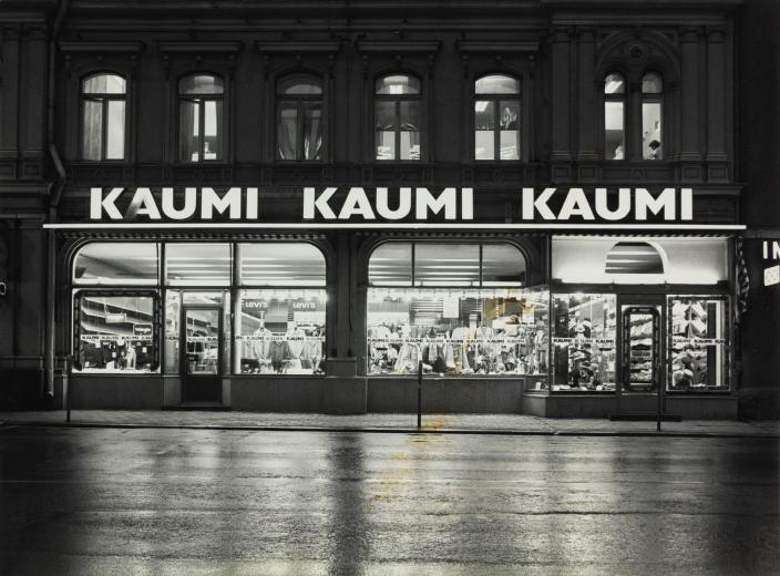 Tunnelmallinen iltakuva Kaumin miesten asusteliikkeestä, valaistu myynti-ikkuna ja yläpuolella valomainoskyltti tekstillä Kaumi. (Kuva: Valokuvaamo Kurki &amp; Kangas / Reprokuva: Eija Karnisto, TKM)