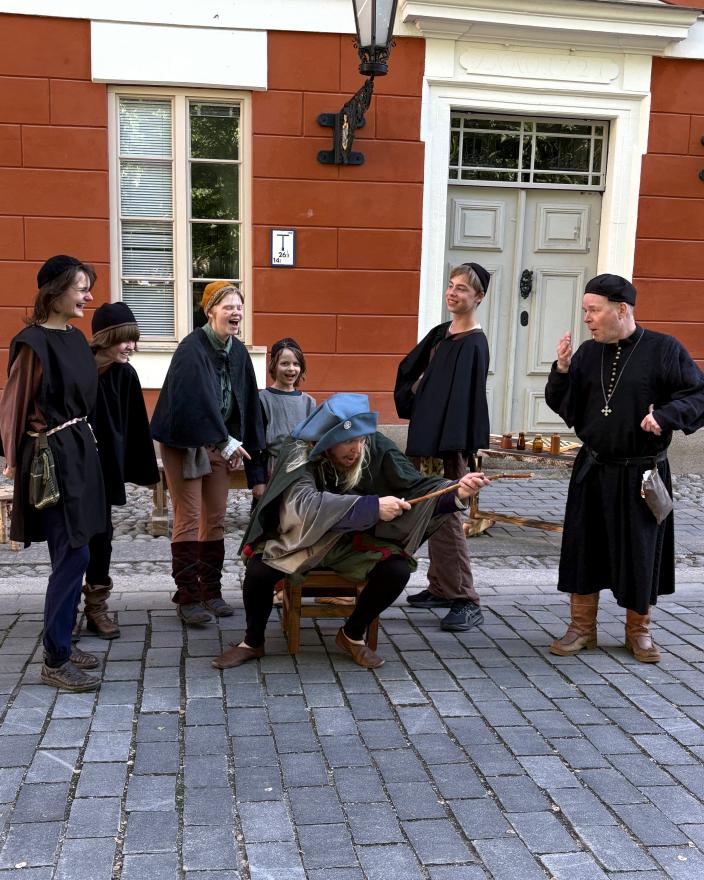 En scen ur en medeltida pjäs. Eleverna och prästen skrattar åt alkemisten som sitter på bänken. 