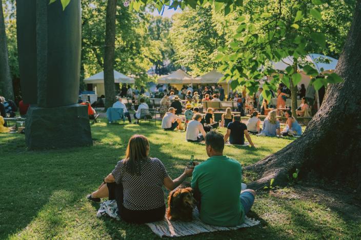 Kaksi ihmistä istuu viltillä koiran kanssa puiden alla, taustalla kojuja ja ihmisiä.
