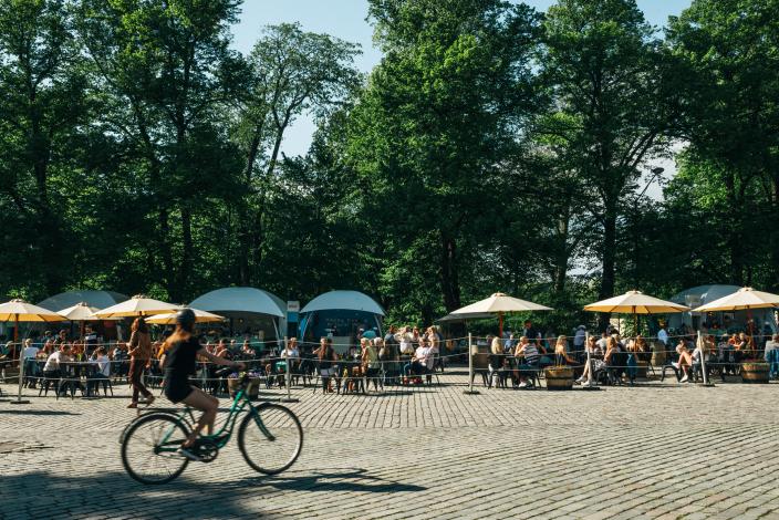 Ihmisiä nauttimassa auringosta ja ruoasta ulkoterassilla suurten puiden katveessa kesäisessä Kirkkopuistossa. Etualalla henkilö ajaa polkupyörällä mukulakivikadulla. (Kuva: Kirkkopuiston Terassi)