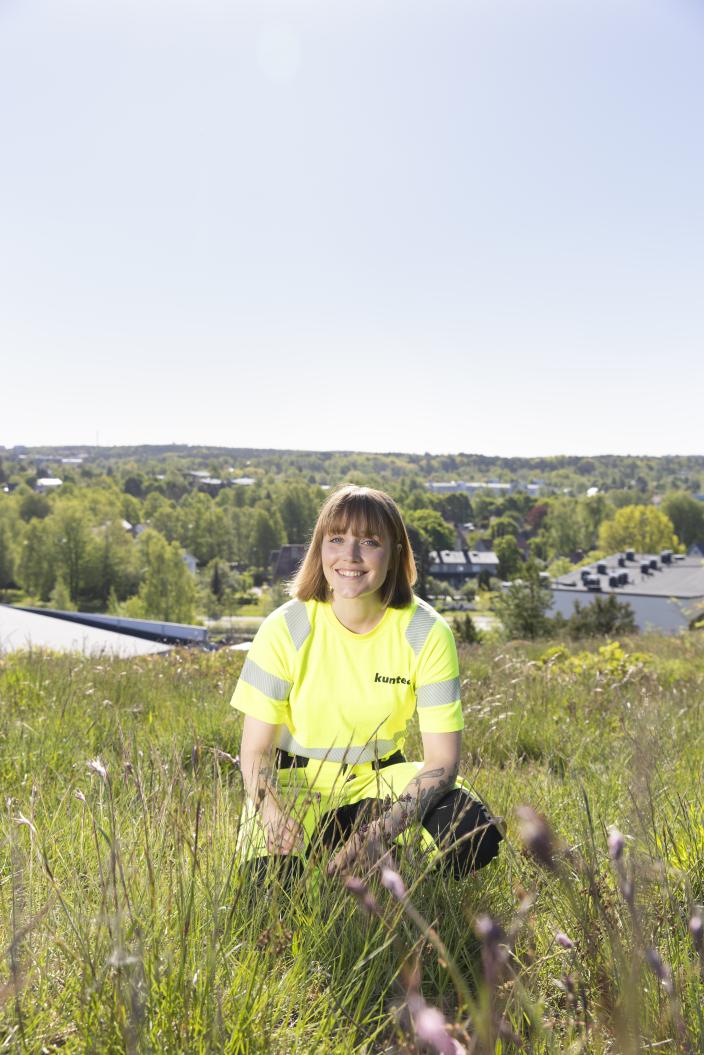 Keltaiseen työasuun pukeutunut henkilö polvistuneena niityllä. Taustalla siintää puustoa ja taloja. (Kuva: Suvi Elo)