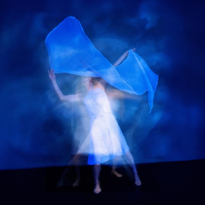 Valkoasuinen henkilö tanssii estradilla sinisävyisessä valaistuksessa. (Kuva: Kulttuuripankki)