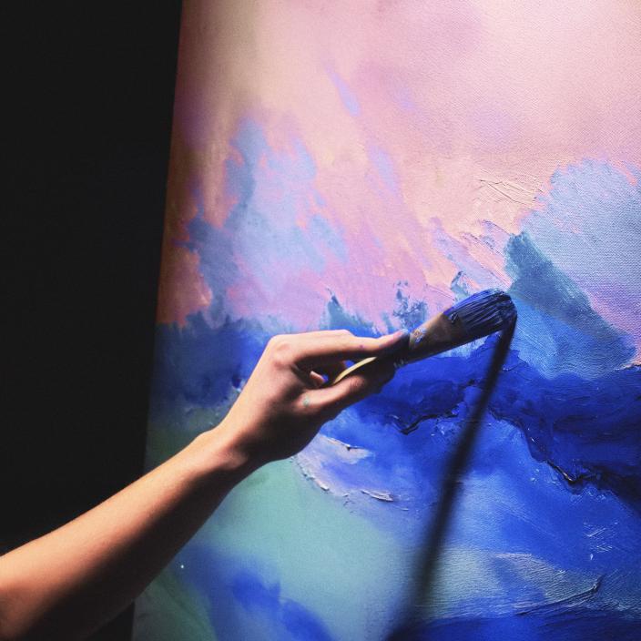 Käsi pitelemässä sivellintä värikkään seinän edessä.