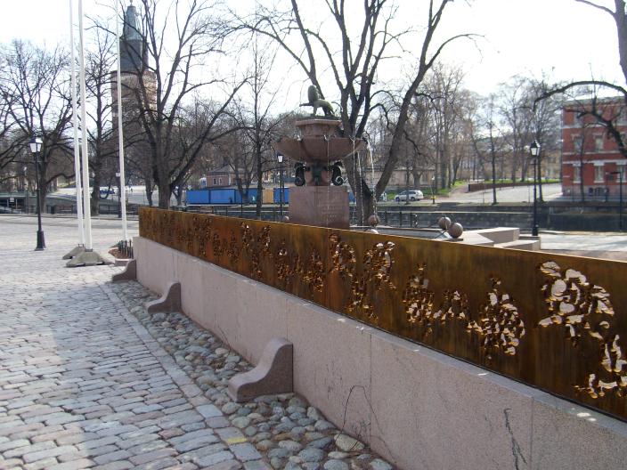 Ruosteenruskea teräksinen taideteos, jota ruusukuviot puhkaisee. Taustalla Leijona-suihkukaivo. (Kuva: Tatiaana Tolonen)