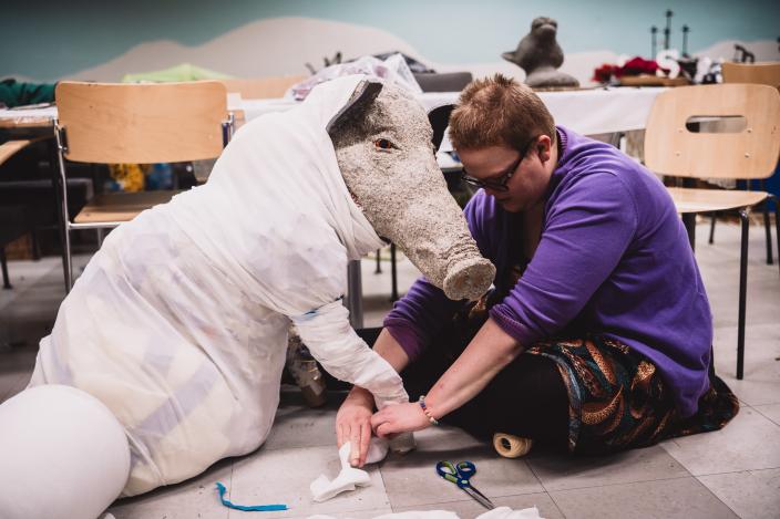 Henkilö istuu lattialla rakentamassa paperimassasta ja kierrätysesineistä villisikaa Aikamatka 20360-teoksessa. (Kuva: Jussi Virkkumaa)