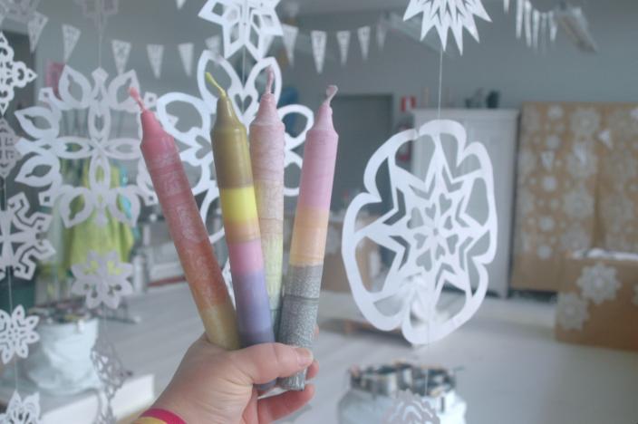 Kuvassa henkilö pitelee neljää värjättyä kynttilää kädessään. Taustalla on huone, jonka katosta roikkuu paperisia lumihiutaleita. (Foto: Hanna Järvenpää)