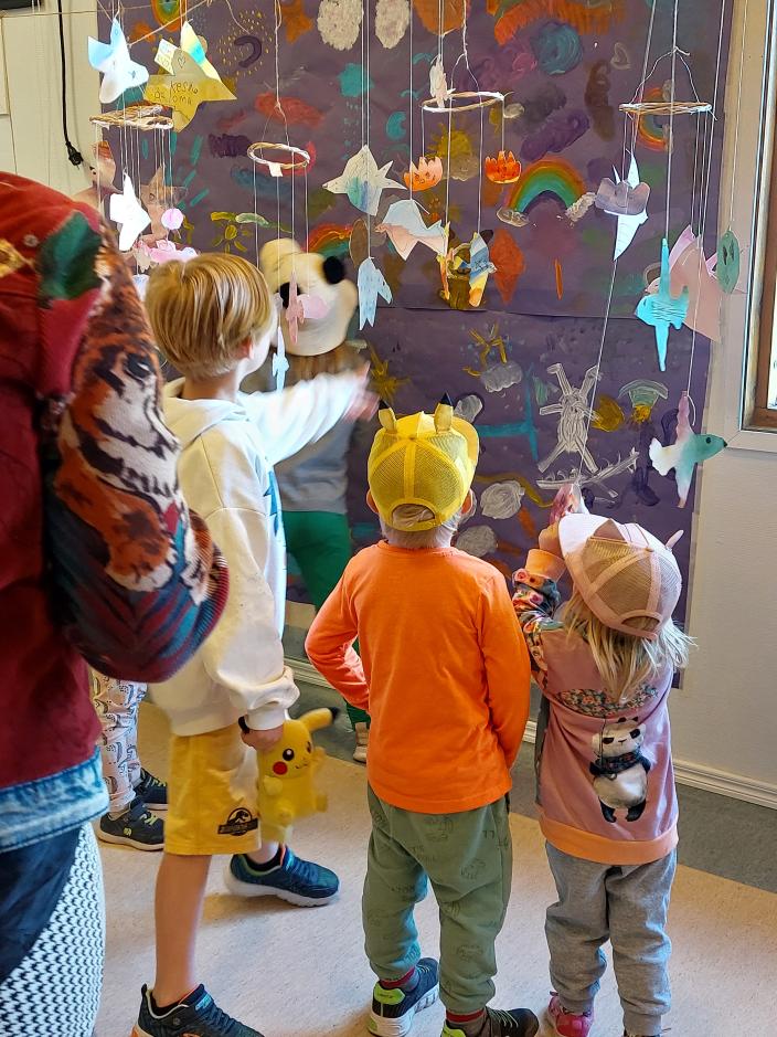 Lapset katselevat suurikokoista maalausta, jonka edessä roikkuu erilaisia paperista askarreltuja lintu- ja eläinhahmoja.  (Kuva: Marika Leinonen-Vainio)