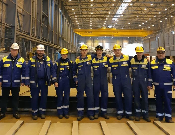 Kahdeksan telakkatyöläistä ja -opiskelijaa seisoo rivissä Meyer Turun telakalla ja poseeraa kameralle.