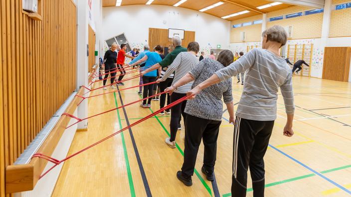 Iso joukko ihmisiä seisoo eri puolilla liikuntasalia selät kameraan päin ja kaikki tekevät samaa kuntoiluliikettä. (Kuva: Timo Jakonen)