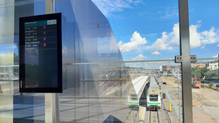 Sähköinen juna-aikataulu Logomon sillalla taustalla rautatiekiskoja ja saapuva juna. (Kuva: Susse Määttänen)