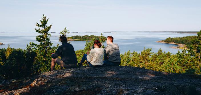 Kolme henkilöä istuskelee korkean kallion laella merimaisemaa katsellen (Kuva: Kim Allen-Mersh)