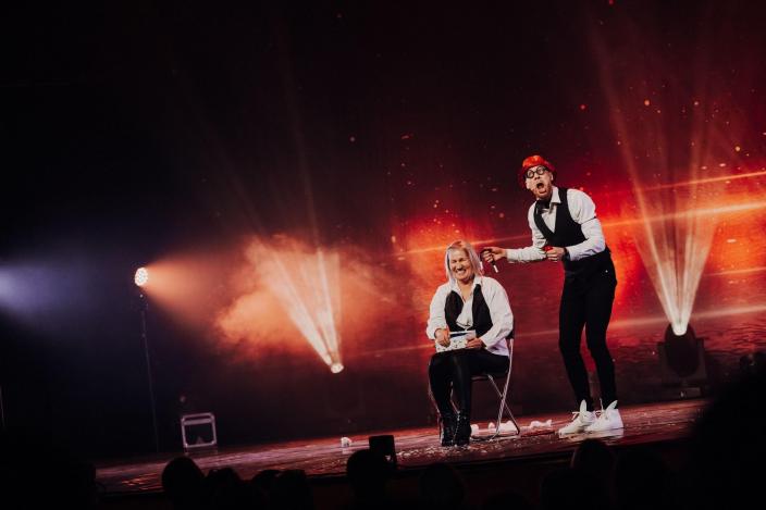 Kuvassa on lavalla naurava yleisöstä valittu avustaja ja taikuri. Tausta on valaistu punaisilla valoilla. (Kuva: Camilla Hanhirova)