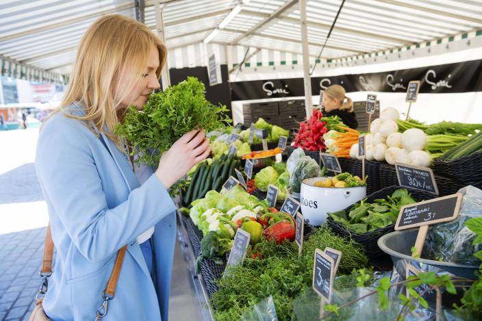 Vaaleatukkainen nainen nuuhkii yrttipuskaa kädessään kauppatorin vihannesmyyjän myyntikojussa. (Kuva: Seilo Ristimäki)