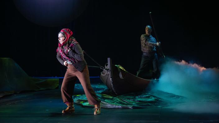 Kaksi näyttelijää esiintyy estradilla Ovllá-oopperassa Oulun teatterissa. Etummainen henkilö vetää köydestä perässään venettä, jossa toinen henkilö seisoo melomassa. (Kuva: Jouni Porsanger)