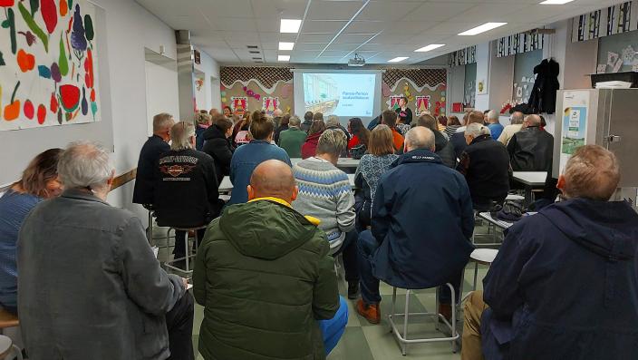 Takaa kuvattu joukko Ihmisiä koulun ruokasalissa kuuntelemassa puhujaa. (Kuva: Päivi Autere)