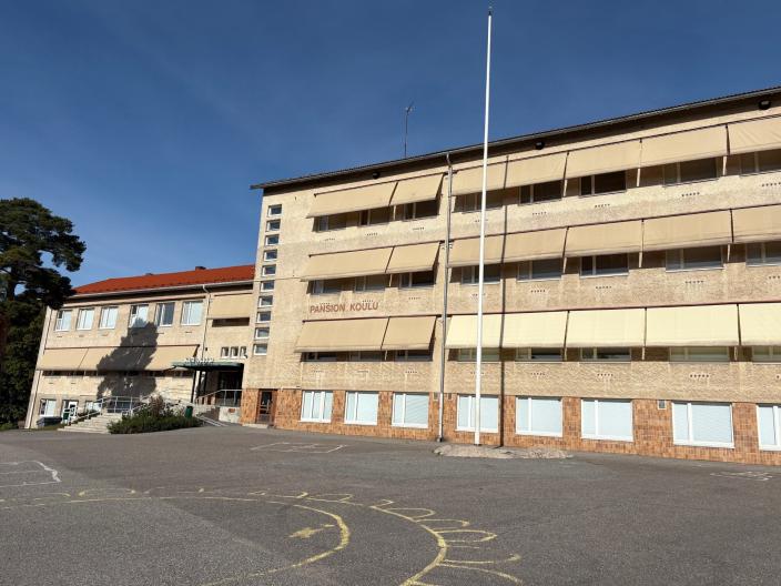 Pansion koulun rakennus koulun pihalta kuvattuna aurinkoisena kesäpäivänä. Rakennuksen edessä lipputanko ja koulunpihan asfaltiin piirretty katuliiduilla kuvioita.  (Kuva: Hannele Sivonen)