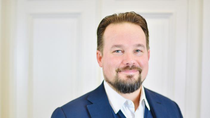 Siniseen pikkutakkiin ja liiviin pukeutunut parrakas henkilö katsoo suoraan kameraan. (Foto: Hannu Oskala / Rud Pedersen Public Affairs)