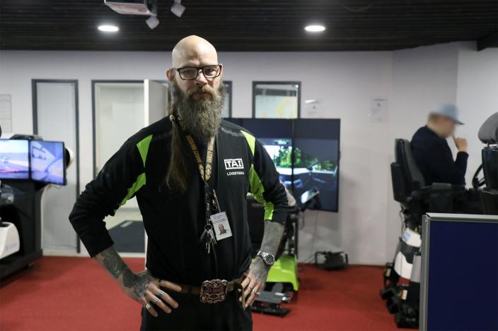 Petri Lapinmäki poseeraa kameralle ajosimulaattoriluokassa. Taustalla näkyy ajosimulaattoria käyttävä opiskelija osittain. (Kuva: Toni Puurtinen)